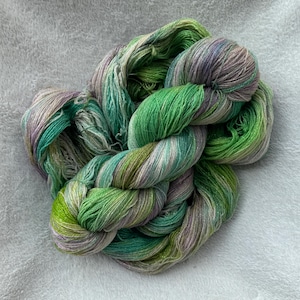 May include: A skein of hand-dyed yarn featuring a variegated color palette of vibrant emerald green, lime green, and soft lavender purple. The fibers are twisted into a loose, textured bundle, showcasing a blend of cool and warm tones throughout.