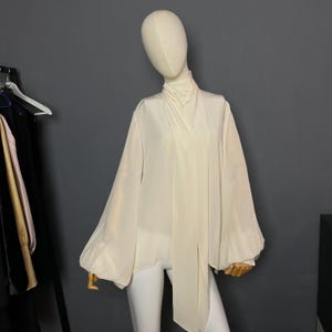 May include: A white, long-sleeved blouse with a high neck and a long tie that hangs down the front. The blouse has a loose, flowing fit and is made of a lightweight fabric.