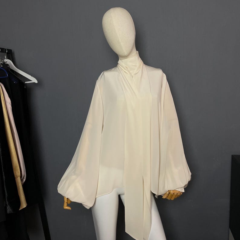 May include: A white, long-sleeved blouse with a high neck and a long tie that hangs down the front. The blouse has a loose, flowing fit and is made of a lightweight fabric.