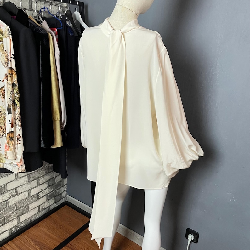 May include: A white blouse with a long, flowing tie at the back. The blouse has long sleeves with a gathered detail at the cuffs.