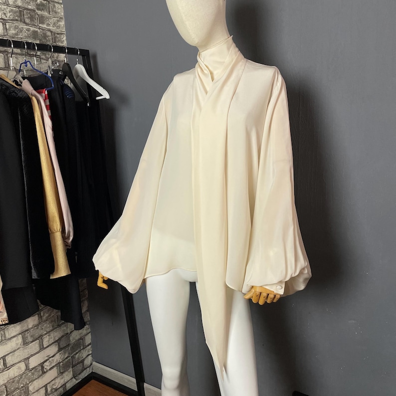 May include: A white, long-sleeved blouse with a high neck and a tie at the front. The blouse has a loose, flowing fit and is made of a lightweight fabric.