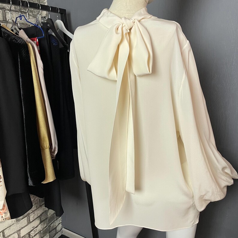 May include: A white long-sleeved blouse with a large bow at the back of the neck. The bow is made of the same fabric as the blouse and is tied in a loose knot.