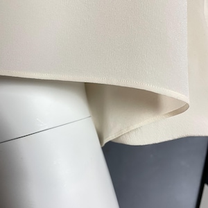 May include: Close-up of a cream-colored fabric with a subtle texture. The fabric has a stitched edge and is folded over.