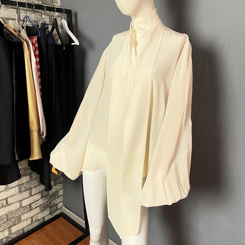 May include: A white, long-sleeved blouse with a high neck and a tie detail at the front. The blouse has a loose, flowing fit and is made of a lightweight fabric.