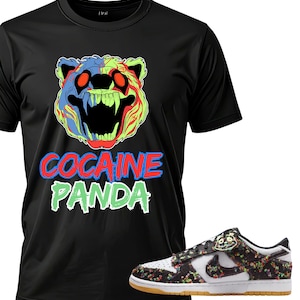 May include: Black t-shirt with a colorful graphic of a bear with the text "COCAINE PANDA" in red and green. A pair of floral print sneakers with a white base and brown sole are next to the shirt.