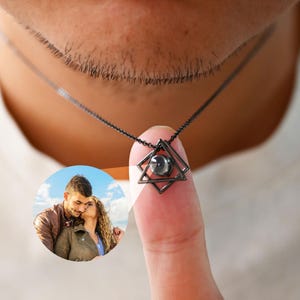 May include: A black geometric pendant necklace with a small, round projection. The pendant is a star shape within a square. The necklace is worn by a person wearing a white shirt. A small, circular image of a couple is visible.
