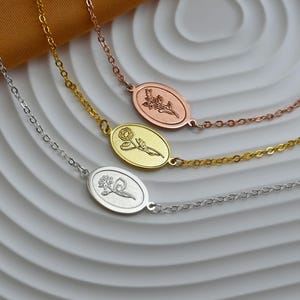 May include: Three delicate bracelets in silver, gold, and rose gold. Each bracelet features a chain and an oval pendant with a floral design. The bracelets are displayed on a white surface with a gold-colored fabric in the background.