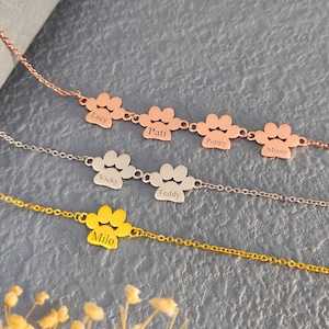 Puede incluir: Tres collares de plata, oro y oro rosa con dijes en forma de pata. Cada dije está grabado con un nombre diferente: Lucy, Patti, Poppy, Missy, Vicky, Teddy y Milo.