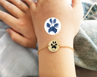 May include: Gold bracelet with a round charm featuring a black paw print and the name "OzZy" engraved on it. The bracelet is on a person's wrist.