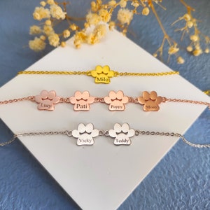 May include: Five personalised pet paw print bracelets with names engraved on each charm. The bracelets are in gold, rose gold, and silver.