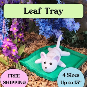 May include: A green leaf-shaped tray with a white ceramic mouse figurine on top. The tray is made of a textured material and has a raised edge. The mouse has black eyes and a pink nose. The tray is surrounded by artificial flowers and wood chips. The text "Leaf Tray" is displayed in a green box above the tray. The text "FREE SHIPPING" is displayed in a pink circle below the tray. The text "4 Sizes Up to 13" is displayed in a white box to the right of the tray.