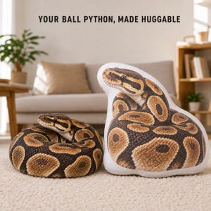 May include: A plush pillow shaped like a coiled ball python with a realistic brown and black scale pattern. The text 'YOUR BALL PYTHON, MADE HUGGABLE' is printed above the pillow. The snake has a detailed head, dark eyes, and a light underbelly.