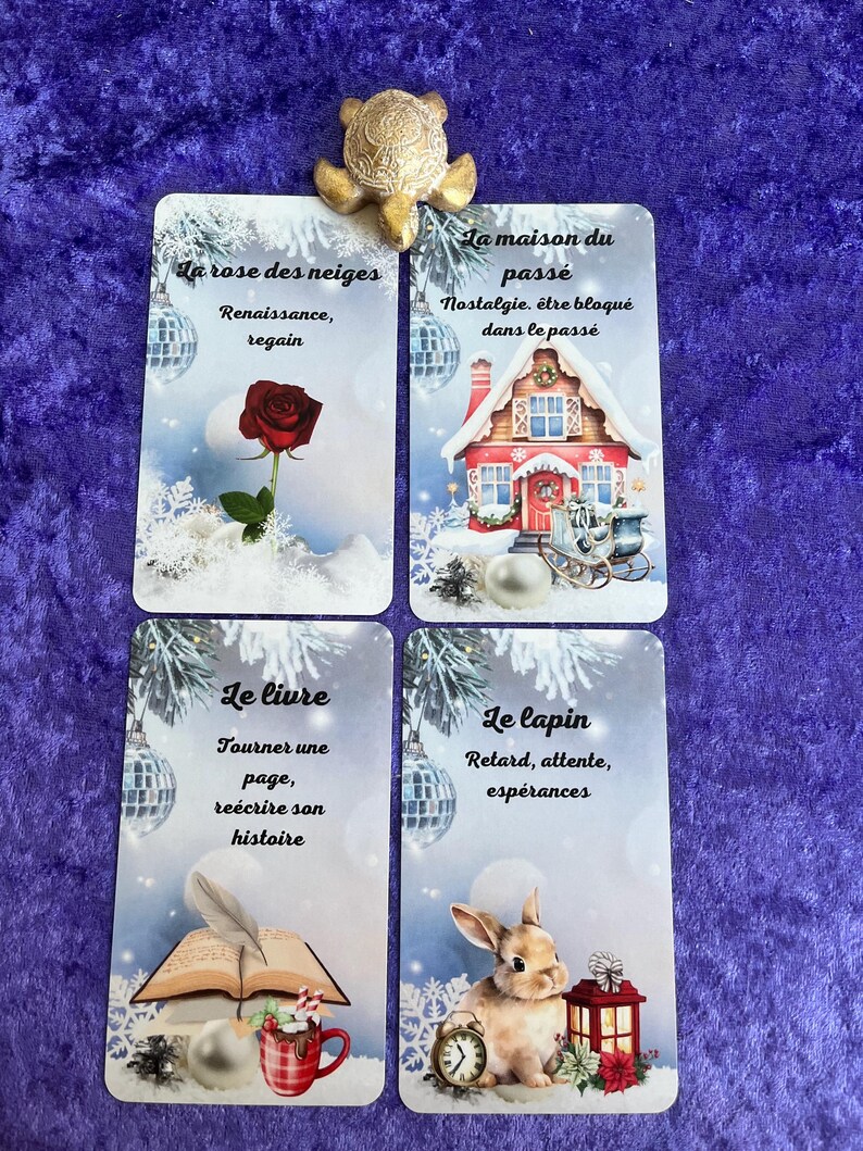 May include: Four tarot cards with winter-themed illustrations and French text. The cards feature images of a rose, a house, a book, and a rabbit. A gold turtle figurine sits atop the cards on a purple velvet surface.