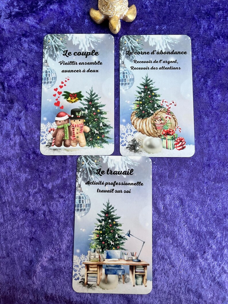 May include: Three tarot cards with winter-themed illustrations. The cards feature images of Christmas trees, ornaments, and text in French. A golden turtle figurine sits above the cards on a purple surface.