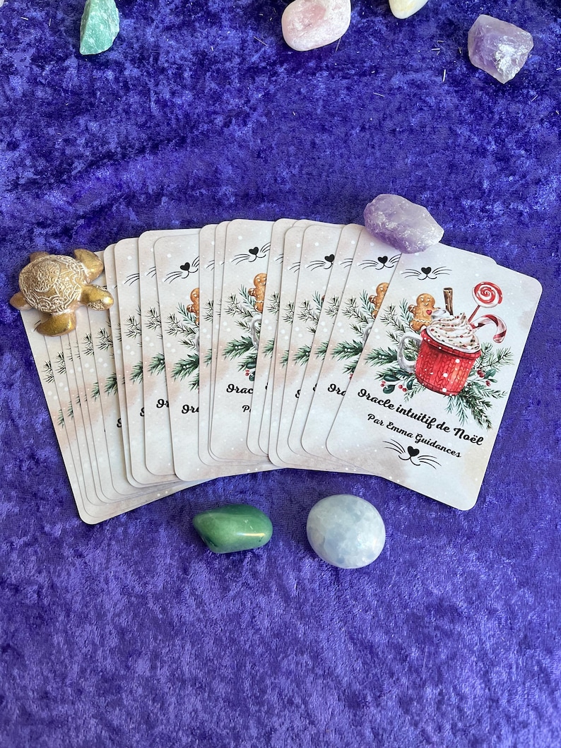 May include: A deck of oracle cards with a Christmas theme, featuring illustrations of gingerbread men, hot cocoa, and festive greenery. The cards are arranged with various gemstones and a decorative turtle figurine on a purple velvet surface.