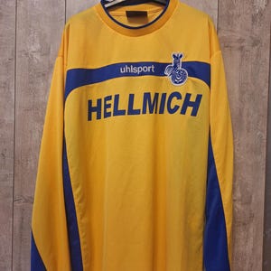 Op de afbeelding: Een gele longsleeve met blauwe accenten en het woord "HELLMICH" in blauwe letters. De shirt heeft een blauwe streep over de borst met het merk "uhlsport" en een logo. De mouwen hebben blauwe panelen.