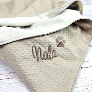 Op de afbeelding: Een beige honden deken met een wafelstructuur en een witte fleece voering. De deken heeft een pootprint en de naam "Nala" geborduurd.