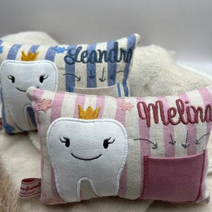 May include: Embroidered tooth fairy pillows. One is pink and white striped with a smiling tooth, a crown, the name "Melina", and a pocket. Another is blue and white striped with the name "Leandro".