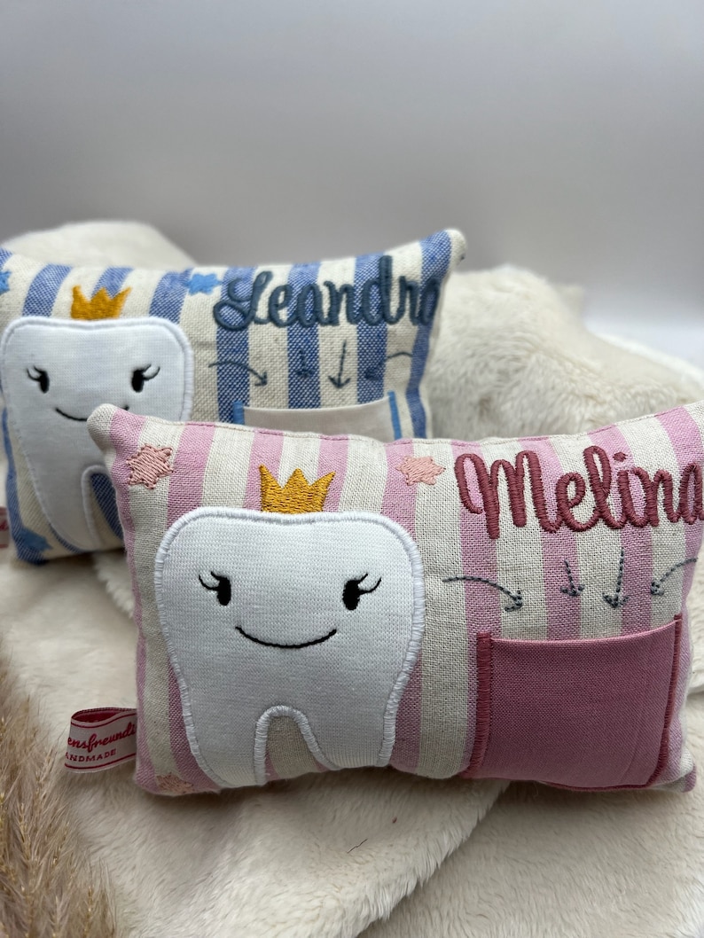 May include: Embroidered tooth fairy pillows. One is pink and white striped with a smiling tooth, a crown, the name "Melina", and a pocket. Another is blue and white striped with the name "Leandro".
