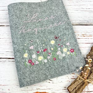 Könnte beinhalten: Eine graue Filzhülle mit gestickten Blumen und dem Text "Linas Zeugnisse" in Rosa. Die Stickerei umfasst Blumen, Schmetterlinge und Grünzeug. Die Hülle wird auf einer weißen Holzoberfläche präsentiert.