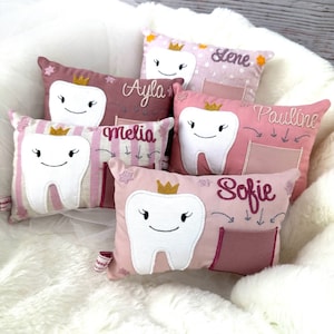 May include: A selection of tooth-shaped cushions in shades of pink and white. Each cushion has a smiling tooth design, a gold crown, and a name embroidered on it. The cushions are displayed on a fluffy white surface.