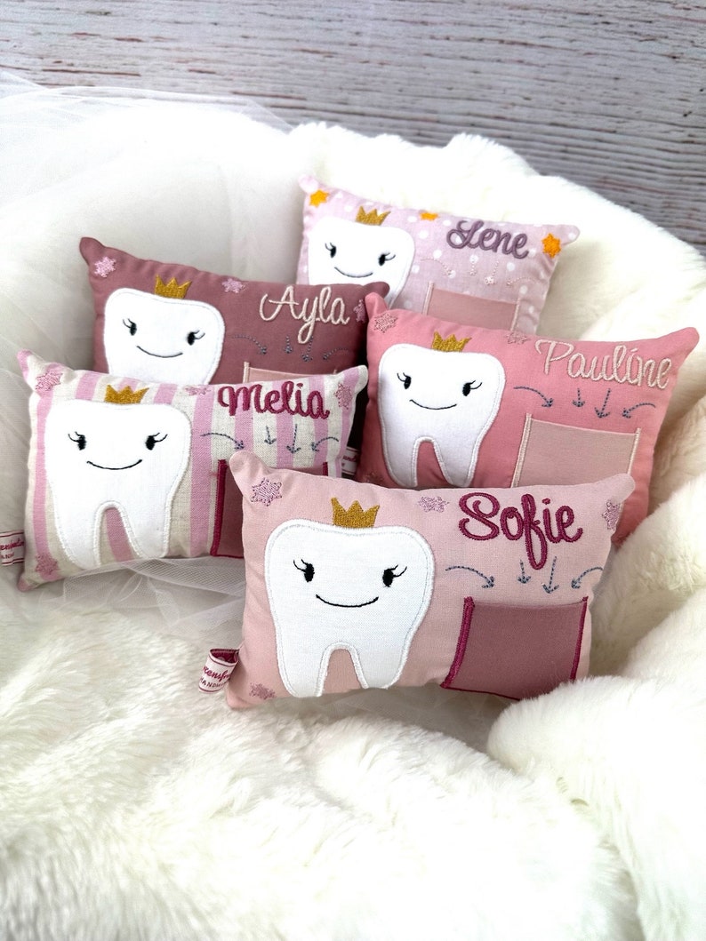 May include: A selection of tooth-shaped cushions in shades of pink and white. Each cushion has a smiling tooth design, a gold crown, and a name embroidered on it. The cushions are displayed on a fluffy white surface.