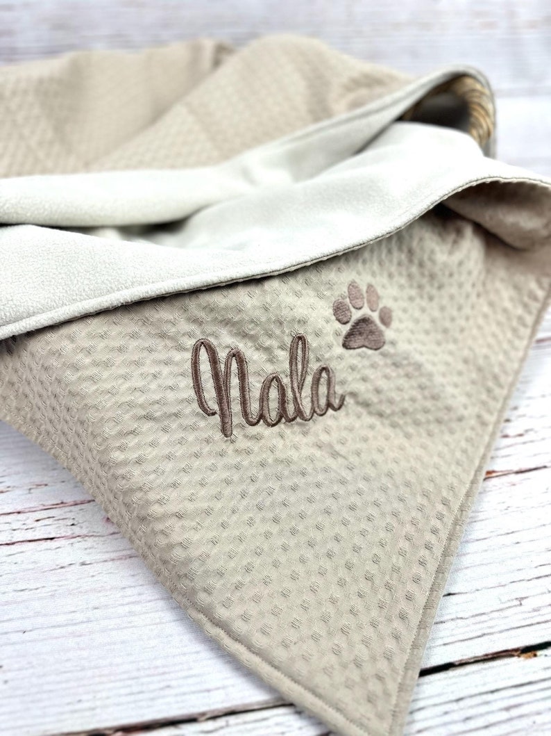 Op de afbeelding: Een beige honden deken met een wafelstructuur en een witte fleece voering. De deken heeft een pootprint en de naam "Nala" geborduurd.