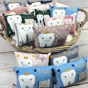May include: A selection of small, tooth-shaped pillows in a wicker basket. Each pillow has an embroidered tooth with a crown and a name. The pillows are in various colours, including blue, pink, and green, with decorative details.