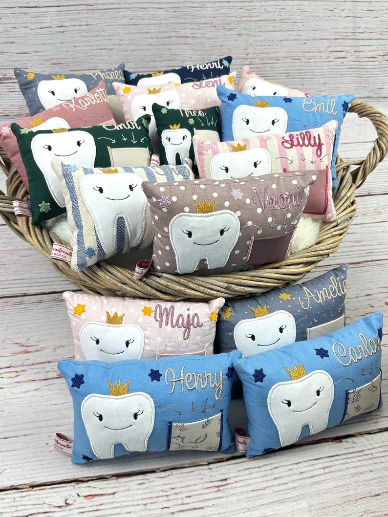 May include: A selection of small, tooth-shaped pillows in a wicker basket. Each pillow has an embroidered tooth with a crown and a name. The pillows are in various colours, including blue, pink, and green, with decorative details.
