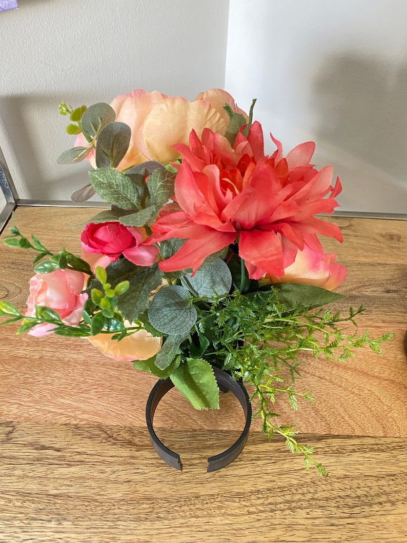 May include: A bouquet of artificial flowers in a black vase. The bouquet includes peach and pink roses, a bright orange dahlia, and green foliage.