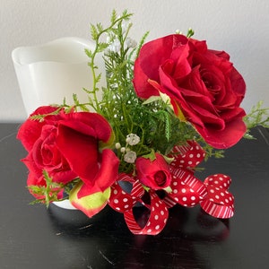May include: A white candle holder with red roses and a red and white polka dot ribbon. The roses are arranged around the candle holder and the ribbon is tied around the base.