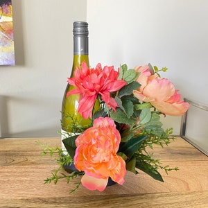 May include: A white wine bottle with a silver cap is placed in a vase with pink and peach flowers and green leaves.