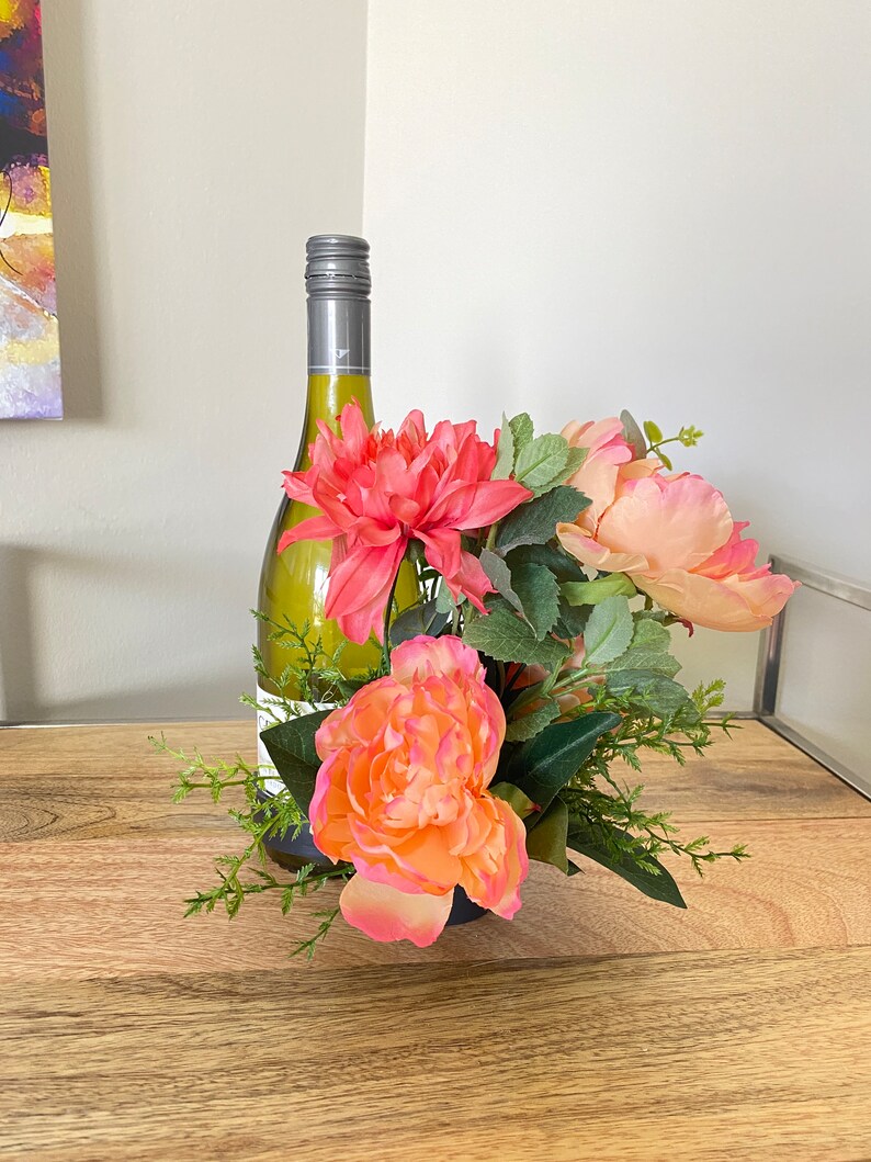 May include: A white wine bottle with a silver cap is placed in a vase with pink and peach flowers and green leaves.