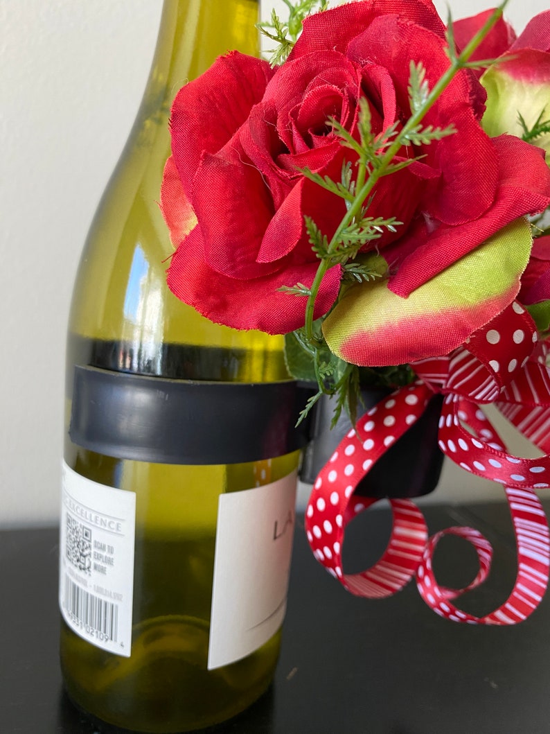May include: A green glass wine bottle with a black ribbon around the neck. A red rose with green leaves is attached to the bottle with a red and white polka dot ribbon.