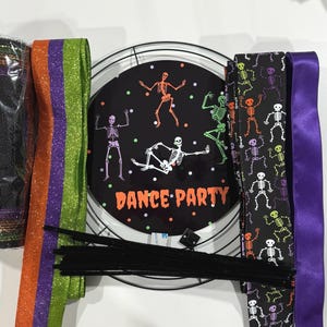 May include: A Halloween wreath kit with a black mesh wreath, orange and purple ribbon, black and white skeleton print ribbon, purple satin ribbon, black pipe cleaners, and a black and white sign that says "Dance Party".
