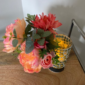 May include: A bouquet of pink and peach artificial flowers arranged in a glass candle holder with a mosaic pattern. The candle holder is lit and the light shines through the glass.