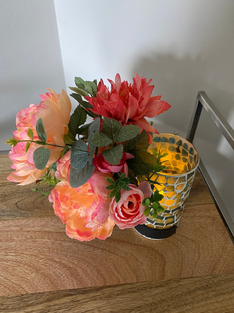 May include: A bouquet of pink and peach artificial flowers arranged in a glass candle holder with a mosaic pattern. The candle holder is lit and the light shines through the glass.