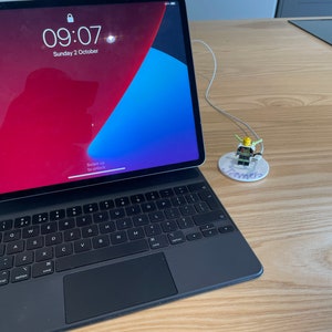 以下が含まれることがあります： 黒いiPadにキーボードが接続され、白い充電ケーブルが接続されています。iPadの画面には、時間09:07と日付10月2日（日曜日）が表示されています。小さなレゴのフィギュアが、「ジェームズ」と書かれた白い円形のスタンドの上に座っています。