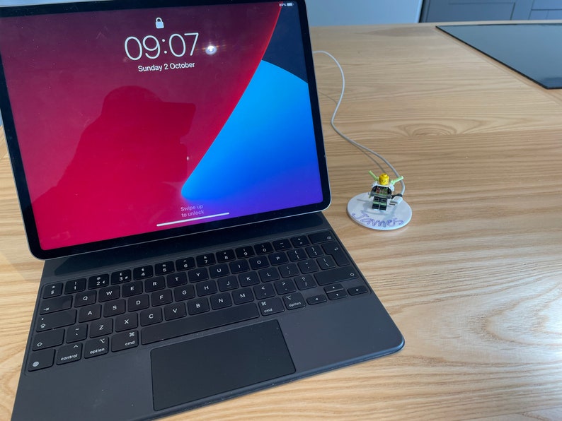 以下が含まれることがあります： 黒いiPadにキーボードが接続され、白い充電ケーブルが接続されています。iPadの画面には、時間09:07と日付10月2日（日曜日）が表示されています。小さなレゴのフィギュアが、「ジェームズ」と書かれた白い円形のスタンドの上に座っています。