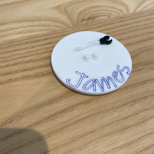 以下が含まれることがあります： 青いインクで「ジェームズ」と書かれた白い丸い名札。タグには、小さな黒と透明なプラスチック製のオブジェクトと、白いボタンが2つあります。