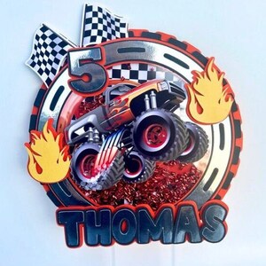 May include: A red, black, and silver monster truck cake topper with a checkered flag, flames, and the number 5. The name "Thomas" is written in black letters below the truck.