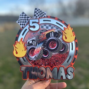 May include: A red, white, and silver monster truck cake topper with a monster truck, flames, checkered flags, and the number 5. The topper also includes the name "THOMAS" in silver letters.