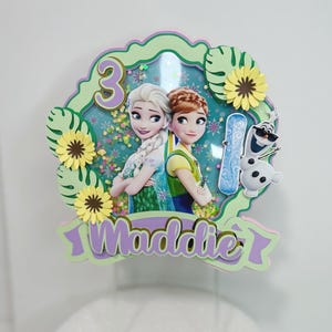 May include: A green, purple, and yellow cake topper with a picture of Elsa and Anna from Frozen. The topper also includes the number 3 and the name Maddie.