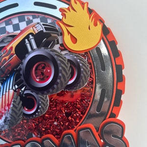 May include: A red, black, and silver monster truck cake topper with a yellow flame and the name "Thomas" in silver letters.