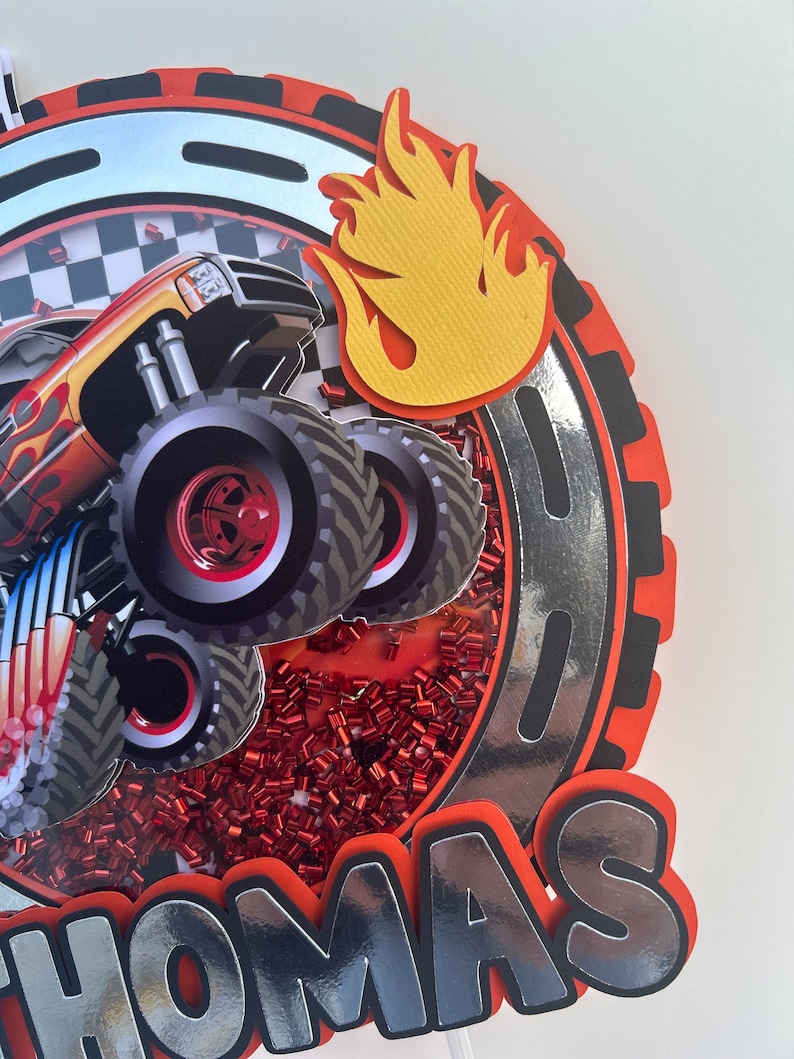May include: A red, black, and silver monster truck cake topper with a yellow flame and the name "Thomas" in silver letters.