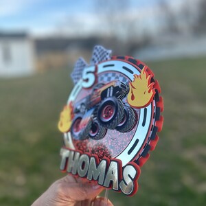May include: A red, black, and silver cake topper with a monster truck design, flames, and the name "Thomas" on it. The topper is on a clear plastic stick.