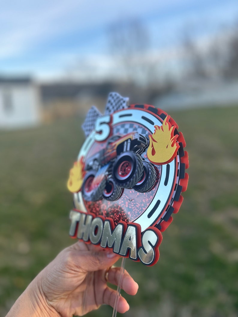 May include: A red, black, and silver cake topper with a monster truck design, flames, and the name "Thomas" on it. The topper is on a clear plastic stick.