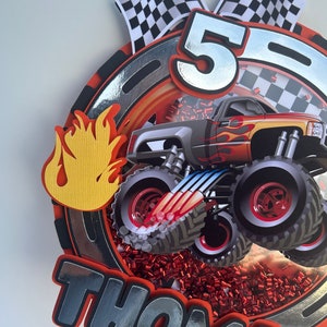 May include: A red, black, and silver monster truck cake topper with a checkered flag, flames, and the name "Thomas" on it. The truck is a monster truck with large tires and a red and yellow flame design. The cake topper is perfect for a monster truck themed birthday party.