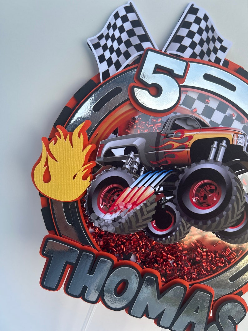 May include: A red, black, and silver monster truck cake topper with a checkered flag, flames, and the name "Thomas" on it. The truck is a monster truck with large tires and a red and yellow flame design. The cake topper is perfect for a monster truck themed birthday party.