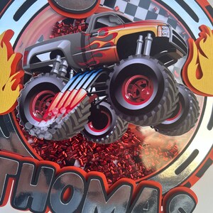 May include: A silver and red monster truck cake topper with the number 5 and the name Thomas. The truck has flames on the side and is surrounded by red confetti.